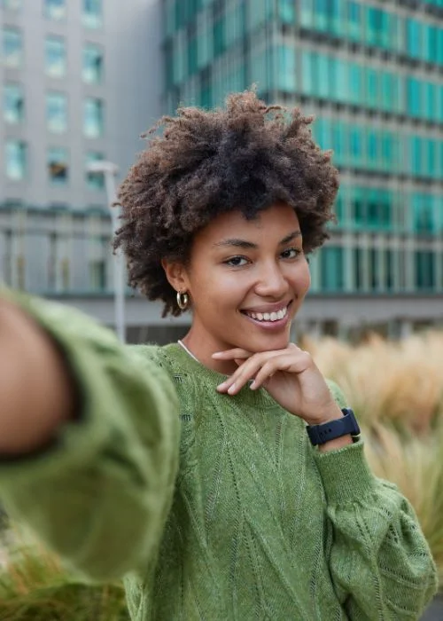 Person smiling confidently at the camera with a hand under the chin, creating a polished and pretty selfie pose.