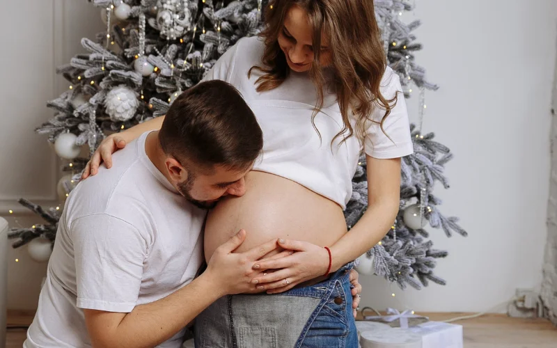 husband kissing the wife's baby bump next to the Christmas tree - Christmas maternity photo ideas