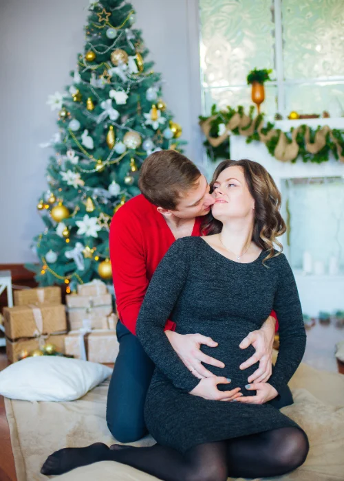 couple posing while kissing next to the Christmas tree - Christmas maternity picture ideas